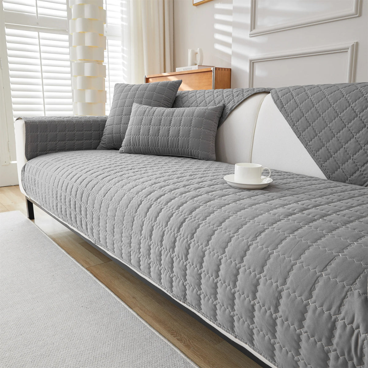 A gray quilted anti-slip sofa cover with white accents and pillows displayed on a modern sofa.