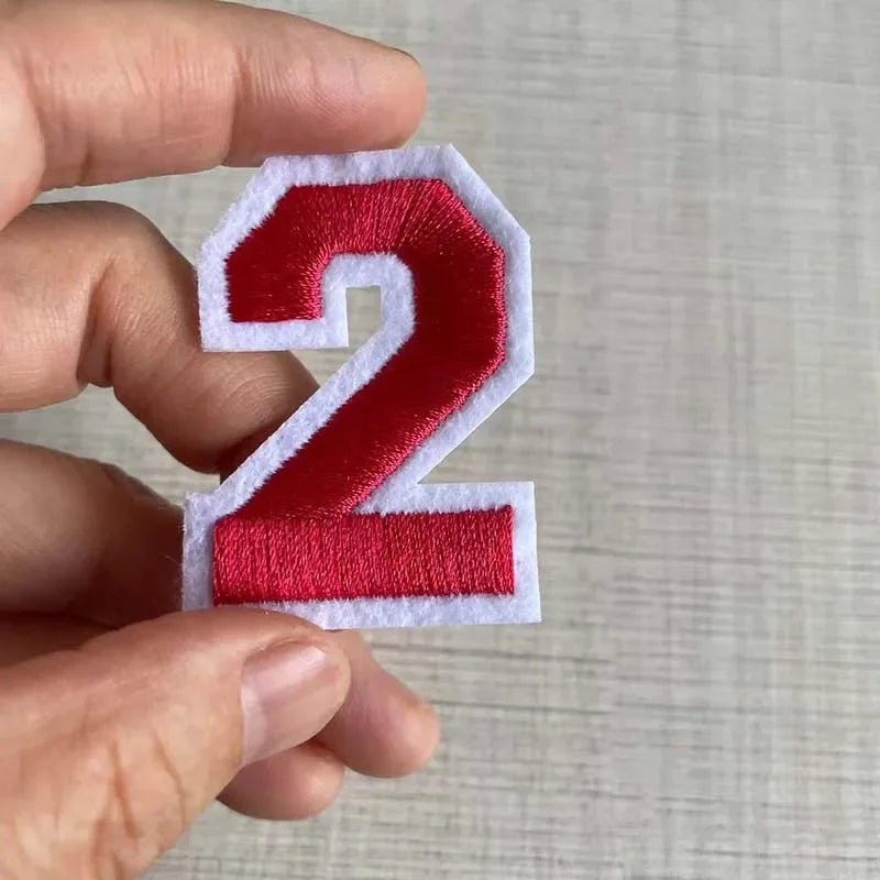 A hand holds a red and white embroidered number 2 patch for apparel.