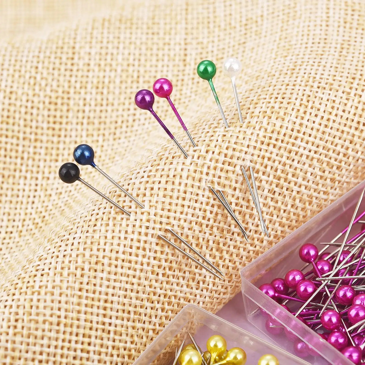 A collection of colorful pearl head sewing pins in various shades are displayed on a burlap fabric.