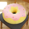 A pink and yellow doughnut-shaped plush pillow with colorful sprinkles resting on a gray fabric surface.