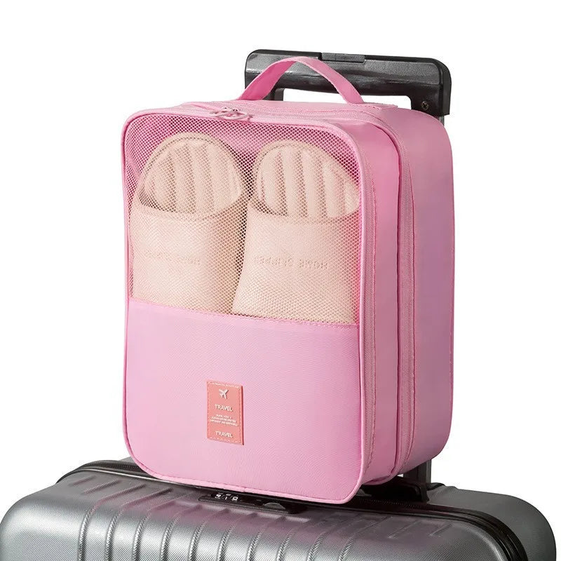 A pink portable travel shoe organizer bag with a mesh pocket and a carrying handle resting on a silver suitcase.