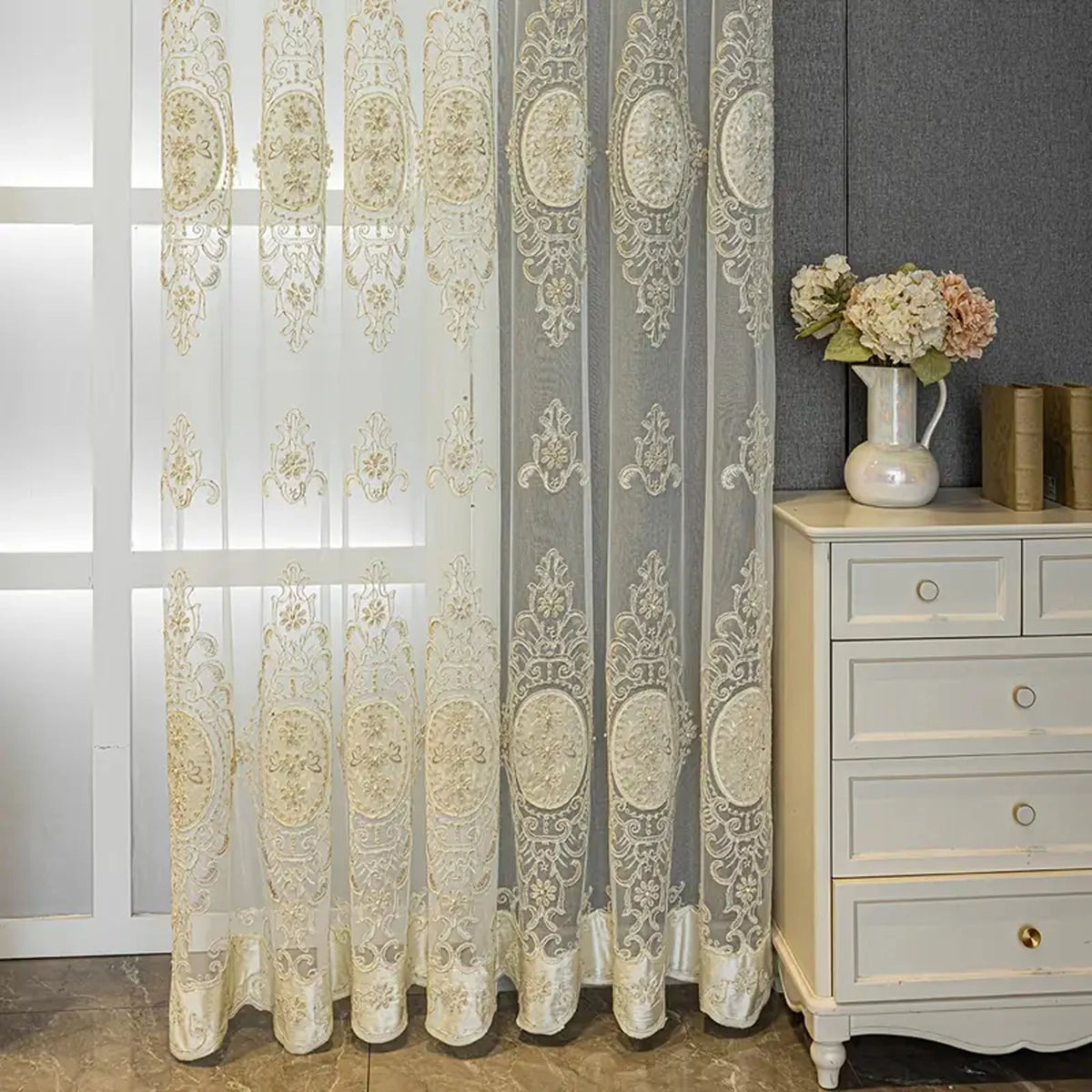 A sheer, embroidered beaded curtain in ivory and gray tones hangs next to a light gray cabinet.