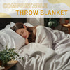A woman is relaxing under a soft, light-colored throw blanket in bed.