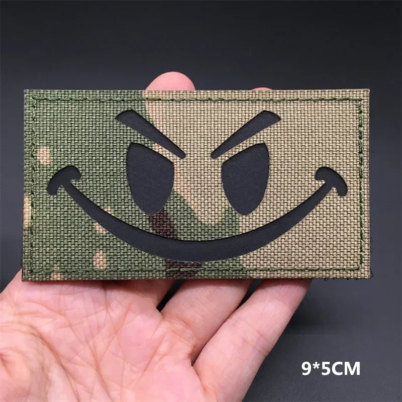 A close-up of a tactical multicam national flag patch with a menacing, angry face design being held in a hand.