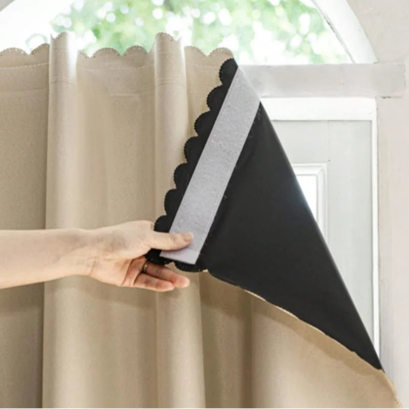 A hand is adjusting a black blackout curtain with a gray hook and loop closure on a light-colored fabric.