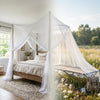 A square mosquito net bed canopy with sheer white fabric draped over a wooden bed in an outdoor setting.