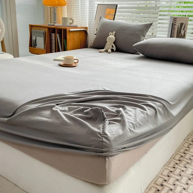 A fitted bed sheet in gray is draped over a bed, with pillows and wooden furniture visible in the background.