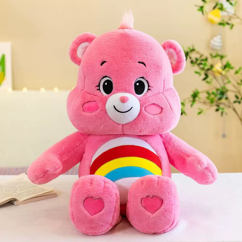 A pink Care Bears Rainbow Bear plush toy with a white belly and heart-shaped paws sitting on a white surface.