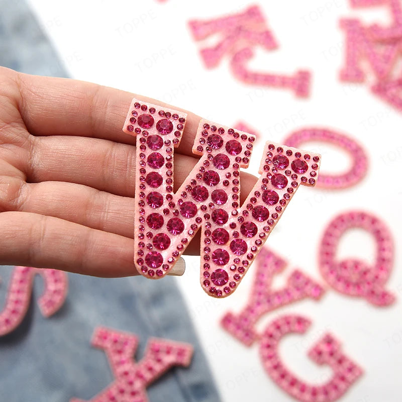A hand holds a pink pearl rhinestone letter patch, part of a set for DIY crafting.