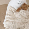 A soft, cream-colored muslin blanket with a floral print and a teddy bear toy is shown.