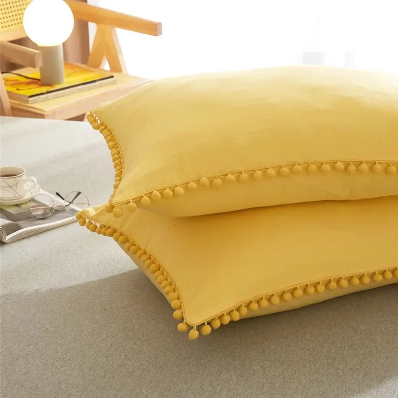 A yellow pillow with pom-pom trim resting on a gray surface.