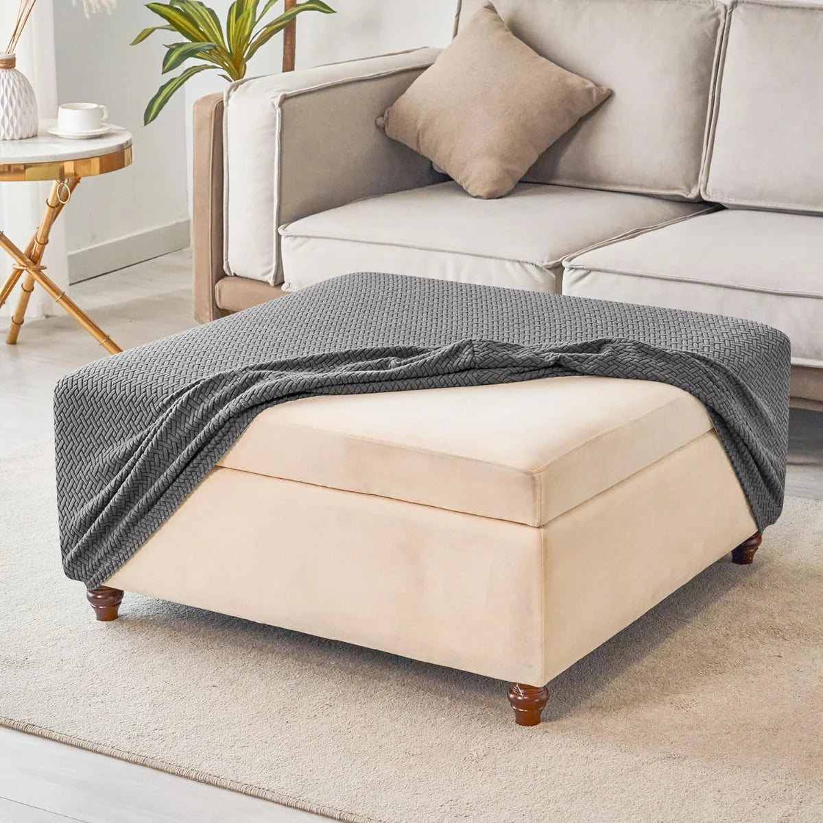 A beige stretch jacquard ottoman and stool cover draped over a light gray sofa in a bright living room.