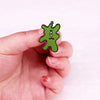 A hand holds a green plankton enamel brooch with a surprised face.