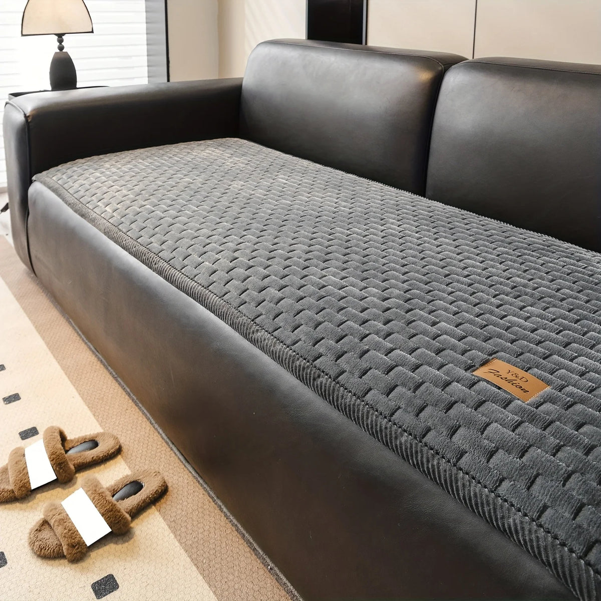 A dark gray plush quilted sofa cover displayed on a black leather sofa with brown slippers nearby.