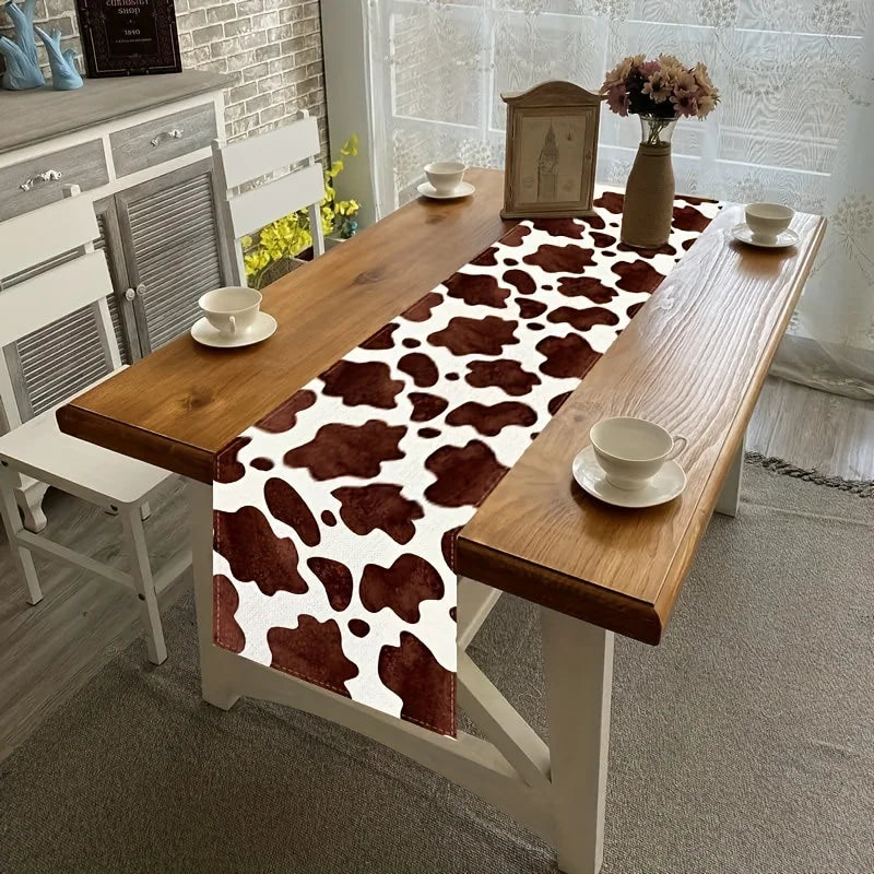 A wooden dining table set with a brown and white cow print linen table runner, cups, and a vase of dried flowers.