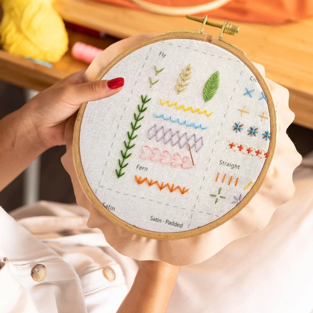 A hand holds a circular embroidery hoop featuring various thread stitches and plant motifs for an adult learning kit.