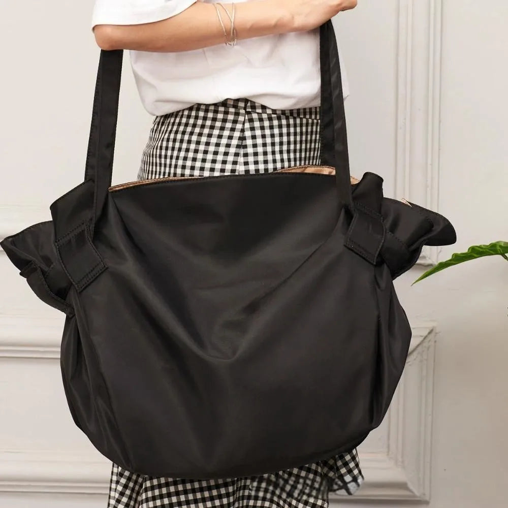 A black foldable tote bag is shown being held by a person, with a plaid skirt visible below.