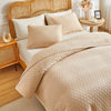 A beige plaid quilt set with quilted covers and pillows on a queen bed with a wooden headboard.