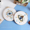 A round embroidery kit featuring a stylized eye design with star patterns on a white fabric against a blue background.