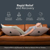 A woman is relaxing on a brown leather sofa using an inflatable leg elevation pillow for muscle relief.