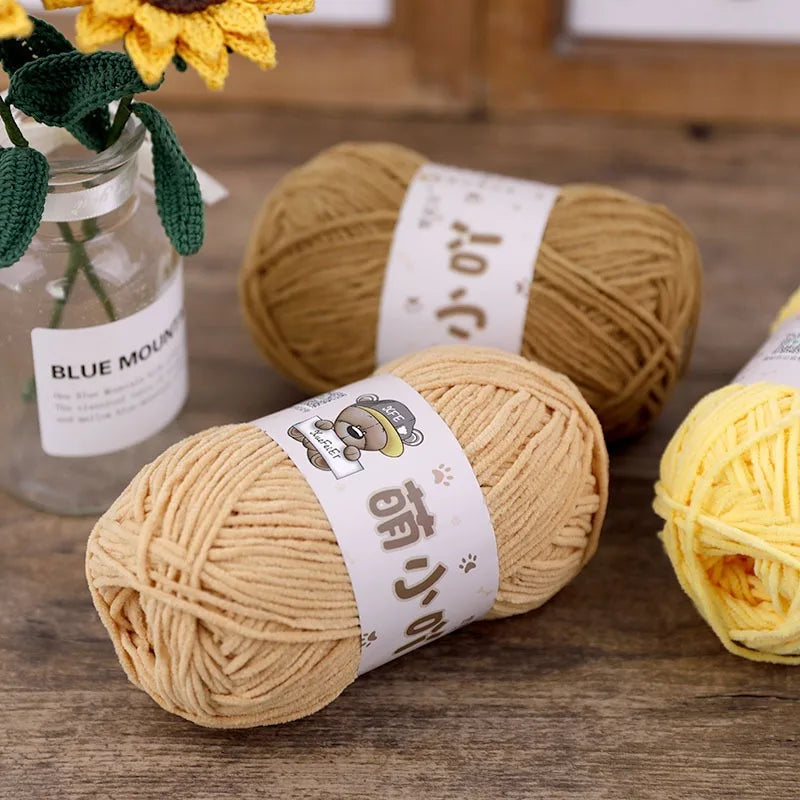 A pile of tan chenille yarn is displayed next to a glass jar and a yellow ball of yarn.