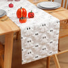 A white table runner with black ghost patterns is placed on a wooden table set for a festive occasion.