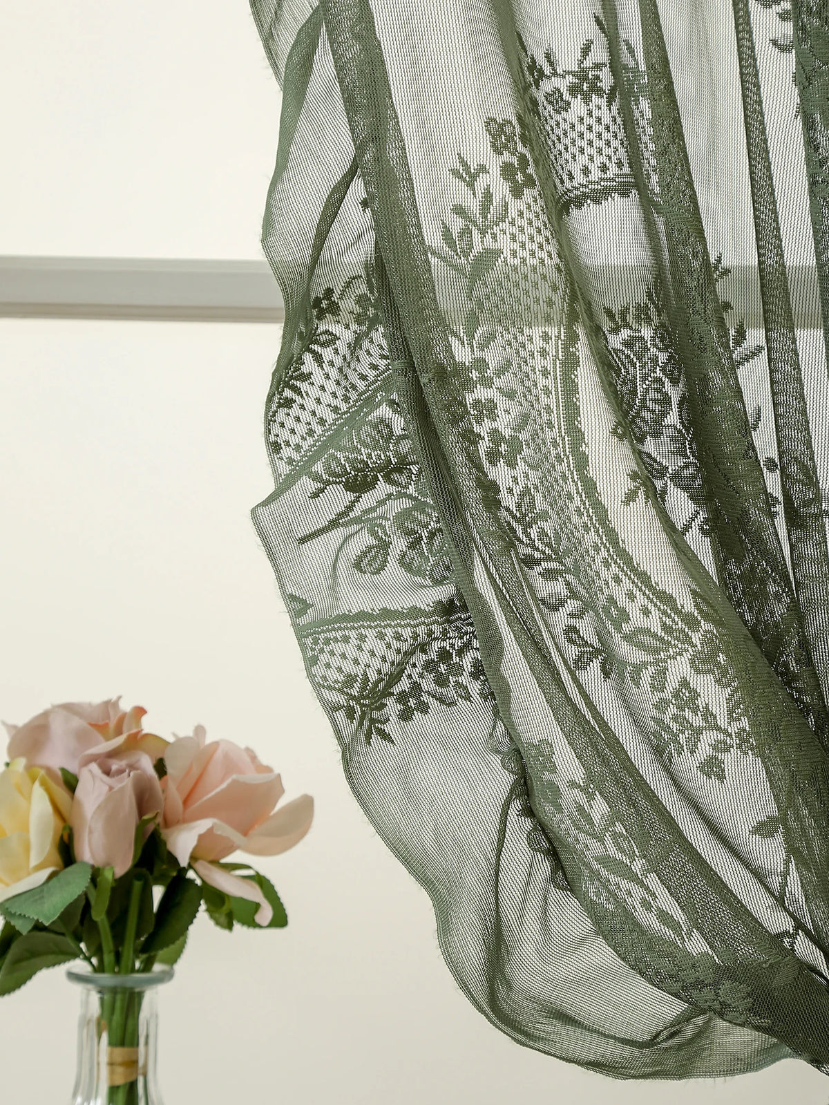 A close-up of sheer moss green lace curtains draped in a window, with a bouquet of pink and yellow roses visible below.