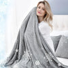 A woman is wrapped in a gray quilted blanket with white snowflake patterns, indoors near a window.