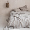 A light gray pure linen flat sheet is draped over a bed, with a woven bag hanging on the wall.