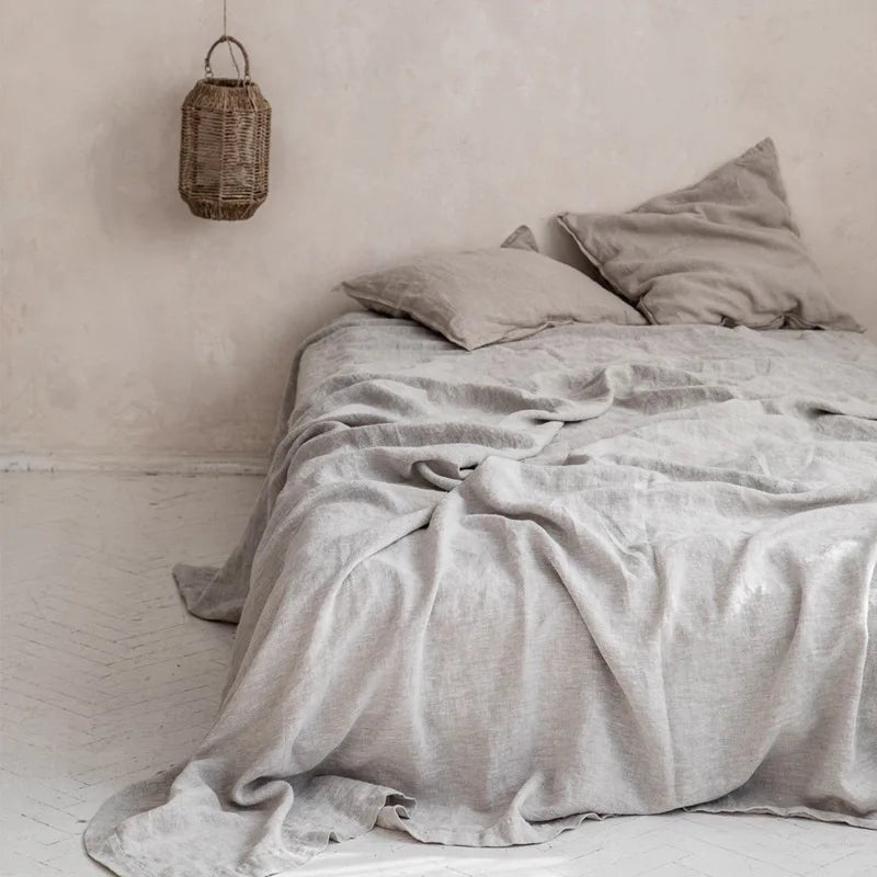 A light gray pure linen flat sheet is draped over a bed, with a woven bag hanging on the wall.