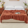 A terracotta colored gauzy blanket is draped over a white bed with white pillows and a book.