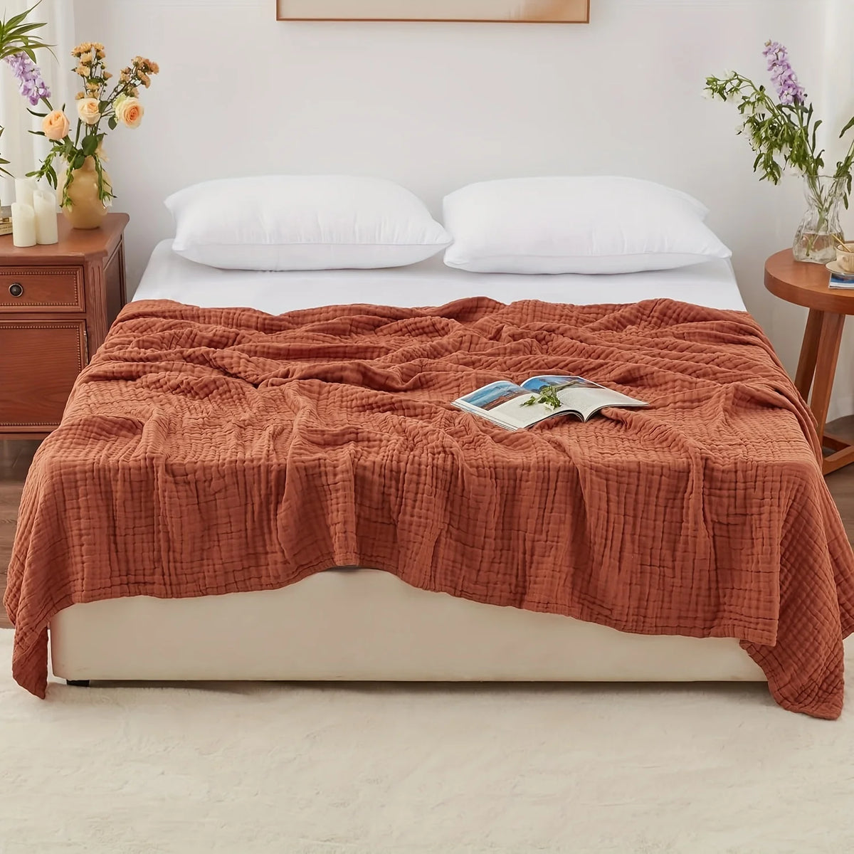 A terracotta colored gauzy blanket is draped over a white bed with white pillows and a book.