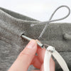 A hand is using a metal tool to thread a spring drawstring onto the opening of a gray knitted garment.