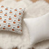 A white throw pillow cover featuring a repeating pattern of small orange ghost and pumpkin designs on a fluffy white rug.
