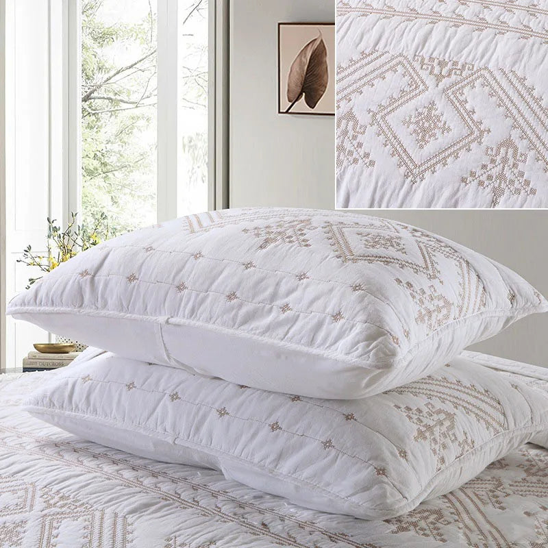 A set of white and beige embroidered cotton quilted pillows resting on a white bed with natural light.