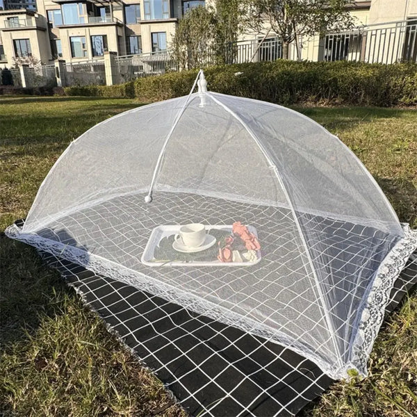 A clear mesh dome tent covering a white teacup and some food items on a black mat outdoors.