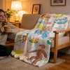 A C-Curious George plush throw blanket draped over a light beige sofa in a brightly lit living room.