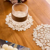 A set of four white lace placemats is displayed on a wooden table next to a glass and some flowers.
