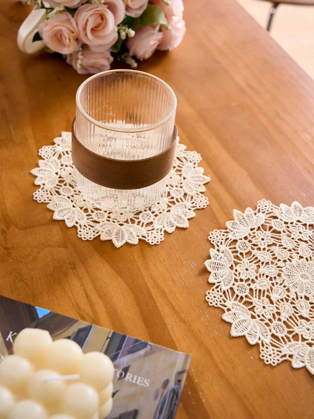 A set of four white lace placemats is displayed on a wooden table next to a glass and some flowers.