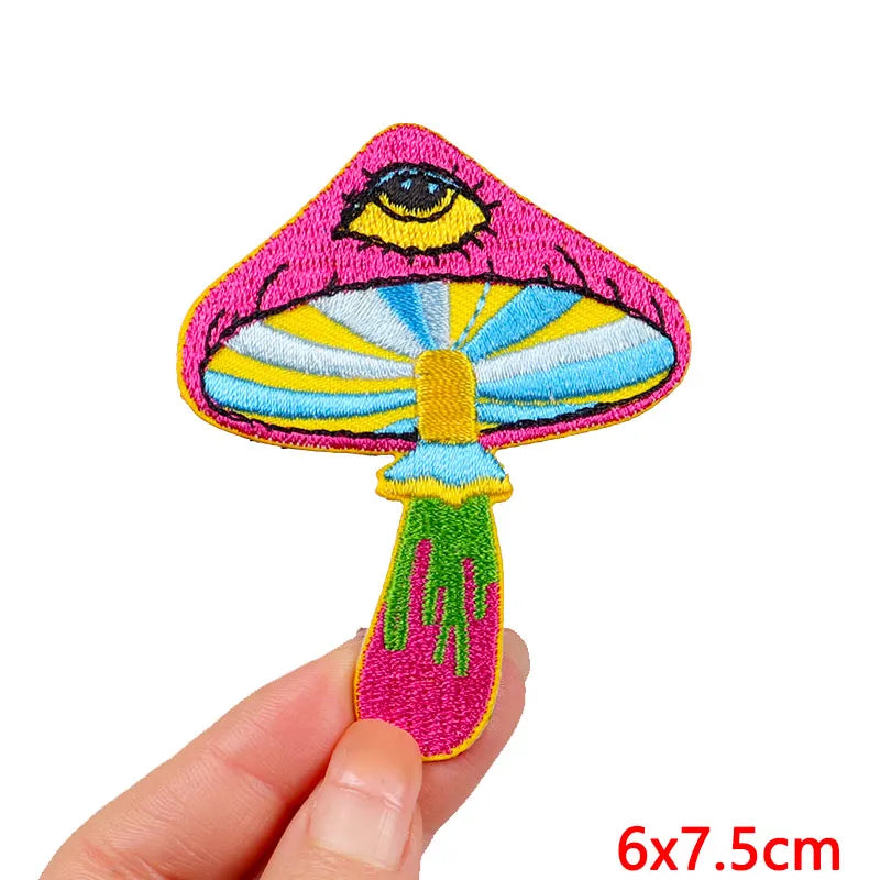 A vibrant pink and yellow embroidered patch shaped like an eye with a tongue, shown being held by a hand.