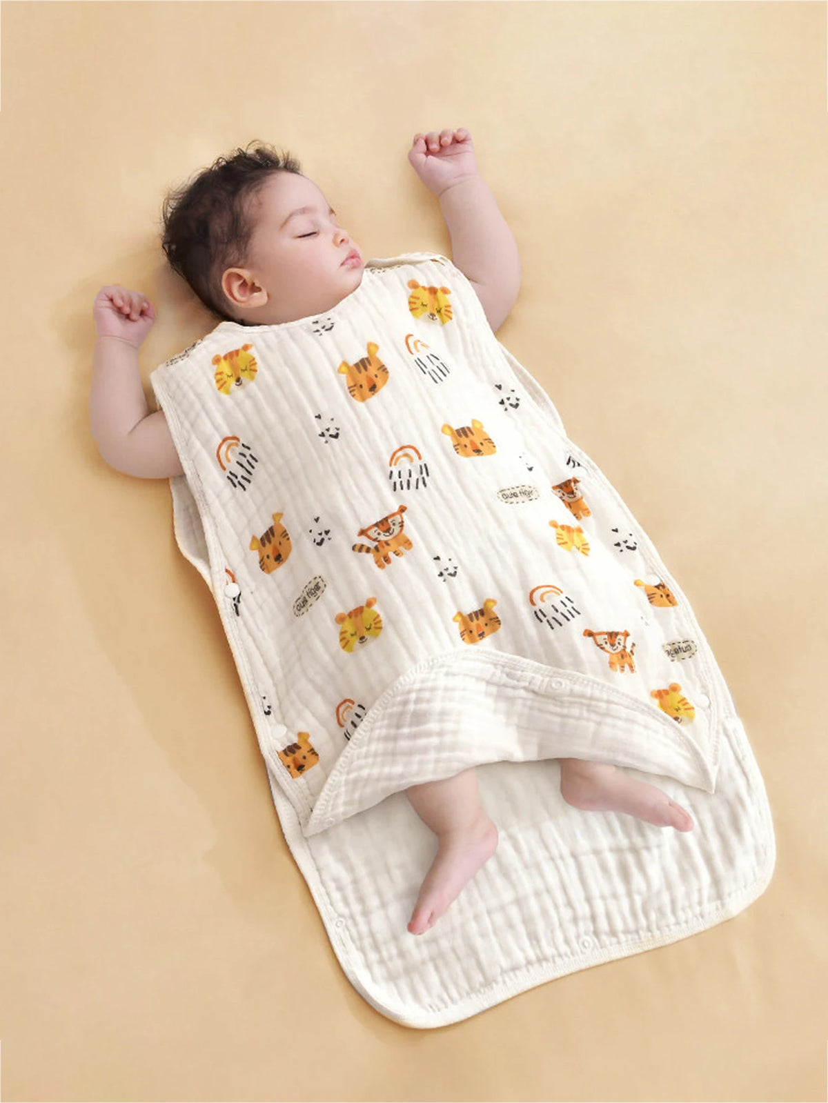 A baby is lying on a beige sleeping bag patterned with cute orange tiger faces and rainbows.