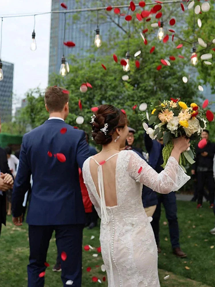 A bride in a white dress is tossing red and white silk rose petals outdoors during her wedding ceremony.