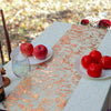 A white table is decorated with an orange sequin glitter runner, red apples, and a glass on an outdoor setting.