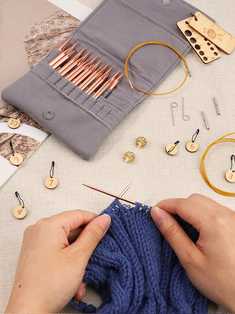 Hands are using an aluminum circular needle set to sew a blue knitted fabric next to a gray storage case.