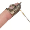 A hand holds an antique sewing thimble and needle protector set with a textured metal thimble and a thin needle.