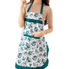 A woman is wearing a floral print kitchen apron with pockets and an adjustable belt over a beige top.