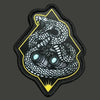 A black military morale patch featuring a detailed silver and yellow depiction of a coiled snake head inside a diamond shape.