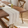 Beige boho hand-woven table runner draped on a dark wood table with a gold glass and wooden chair in the background
