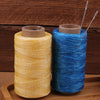 A spool of yellow waxed thread and a blue waxed thread with sewing needles on a wooden surface.