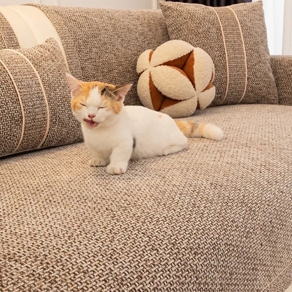 A small orange and white cat is lying on a textured beige sofa cushion surrounded by patterned pillows.